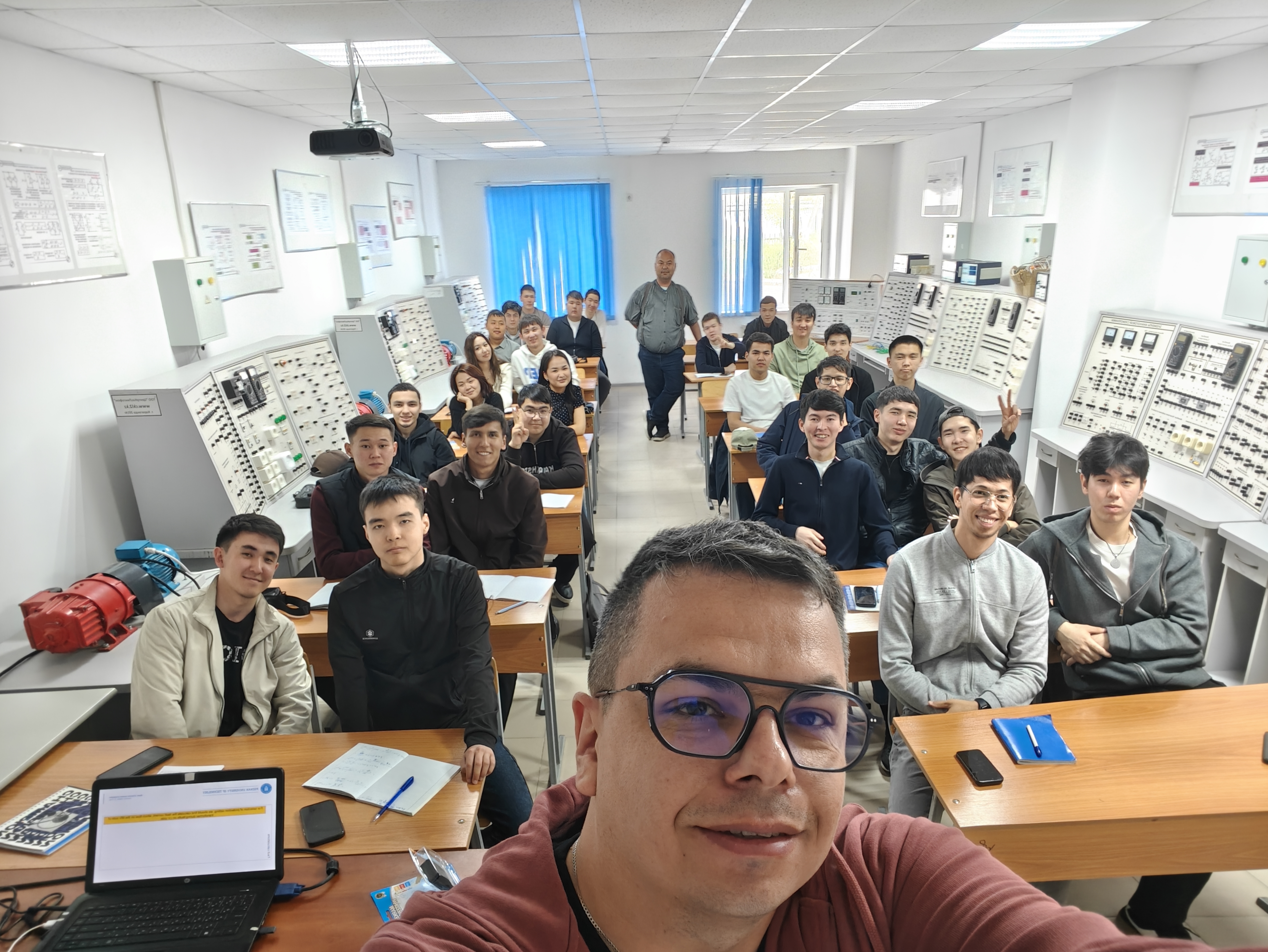 Zdjęcie przedstawiające selfie dr inż. B. Olejnika, w tle którego znajdują się uczestnicy warsztatów. Zdjęcie wykonano w sali laboratoryjnej