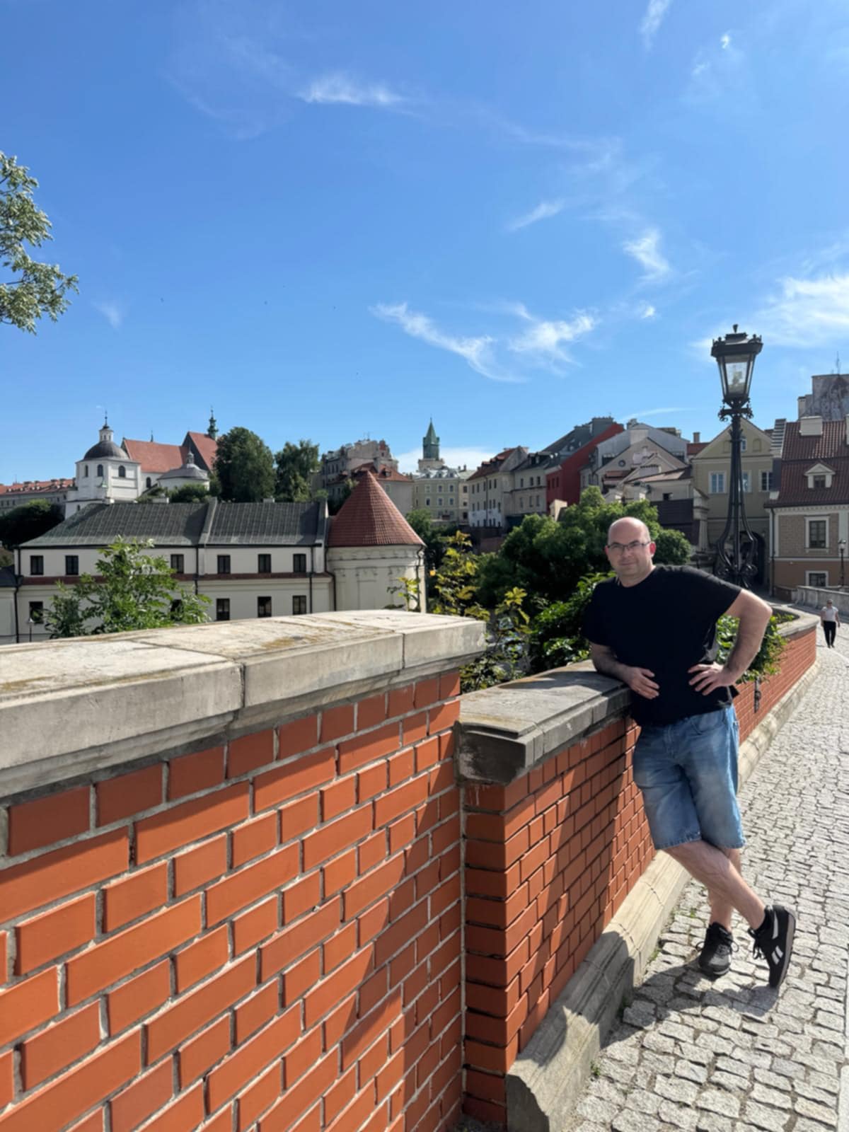 Zdjęcie przedstawia widok na Stare Miasto w Lublinie oraz B.Cerana stojącego przy zabytkowym murze w pobliżu Placu Po Farze