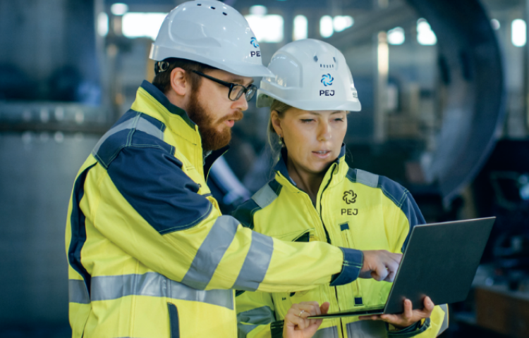Zdjęcie dwojga ludzi w kaskach i odzieży ochronnej, oglądają ekran laptopa. Stoją wewnątrz dużej hali produkcyjnej 