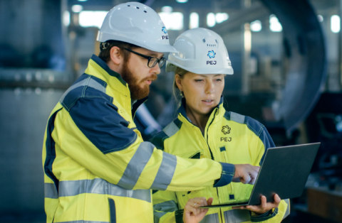 Zdjęcie dwojga ludzi w kaskach i odzieży ochronnej, oglądają ekran laptopa. Stoją wewnątrz dużej hali produkcyjnej 